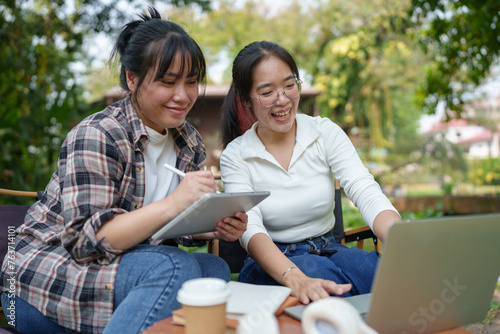 Two Asian female friends laugh and use tablets and laptops to study, learn, and research. Chat Online While relaxing on the grass in the green park holiday concept work lifestyle.