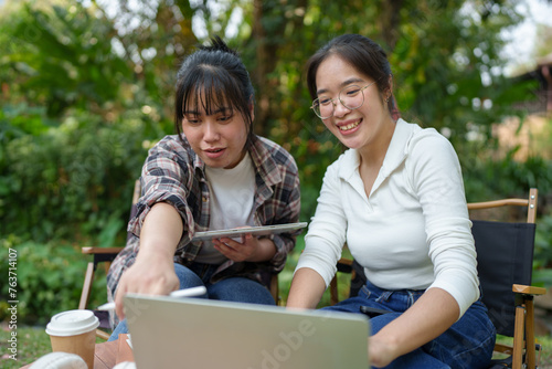 Two Asian female friends laugh and use tablets and laptops to study, learn, and research. Chat Online While relaxing on the grass in the green park holiday concept work lifestyle. © Phimwilai