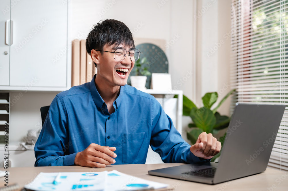 Fototapeta premium job, manager, occupation, freelance, student, corporate, smile, businessman, employee, office. A man is sitting at desk with laptop, stack papers and smiling. He is wearing a blue shirt and glasses.