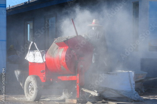 At a construction site, a masonry worker operates an engine-powered mortar mixer with a steel drum, enveloped in a cloud of cement dust as he wears a face mask for protection
