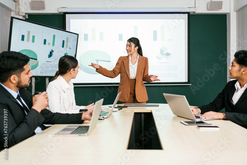 A cheerful and confident Asian businesswoman stands, present bar charts data from projector screen to her office colleagues. Asian business women leader role at the meeting.