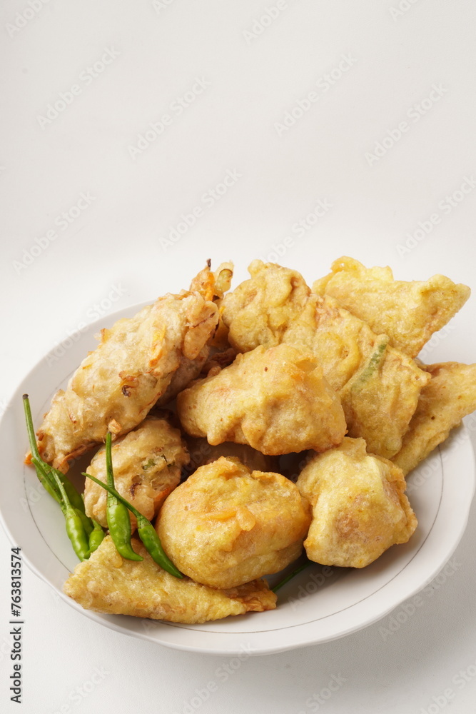 A plate of assorted fried snacks including stuffed tofu, tempeh, and fritters served with bird's eye chili. Captured against a white background. Popular for breaking fast and snacking.