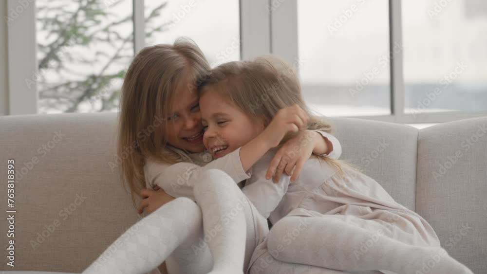 Two children twins playing at home. Happy smiling mischievous little sisters twins sitting on sofa, hugging and laugh, playing in living room at home. Family having fun, sisters' childhood concept. Two children twins playing at home. Happy smiling mischievous little sisters twins sitting on sofa, hugging and laugh, playing in living room at home. Family having fun, sisters' childhood concept.