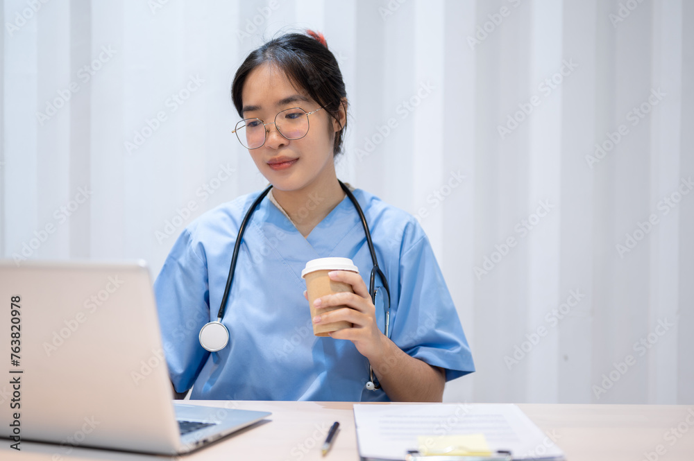 A professional Asian female doctor is sipping coffee while reading medical cases on laptop. A professional Asian female doctor is sipping coffee while reading medical cases on laptop.