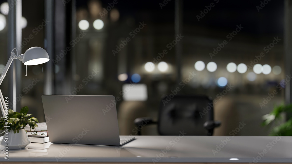 A laptop computer on a desk in a modern private office at night, illuminated by a table lamp. - obrazy, fototapety, plakaty 