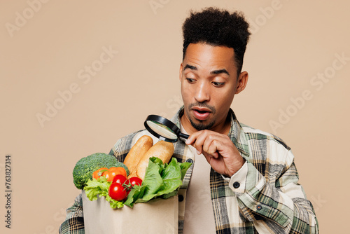 Young shocked man wears grey shirt hold paper bag for takeaway mock up with food products use reading-glass isolated on plain pastel light beige background. Delivery service from shop or restaurant.