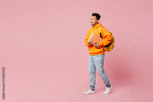 Full body young smart student man of African American ethnicity in yellow hoody casual clothes backpack bag hold books walk go isolated on plain pastel pink background High school university concept