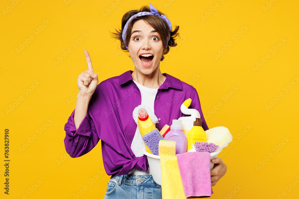 Young woman wear purple shirt hold basin with detergent bottles do housework tidy up hold index finger up with great new idea isolated on plain yellow background studio portrait. Housekeeping concept. Young woman wear purple shirt hold basin with detergent bottles do housework tidy up hold index finger up with great new idea isolated on plain yellow background studio portrait. Housekeeping concept.