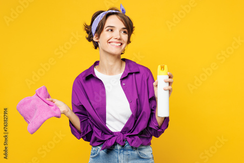 Young woman wears purple shirt casual clothes do housework tidy up hold in hand air freshener spray, rag look aside on area isolated on plain yellow background studio portrait. Housekeeping concept.