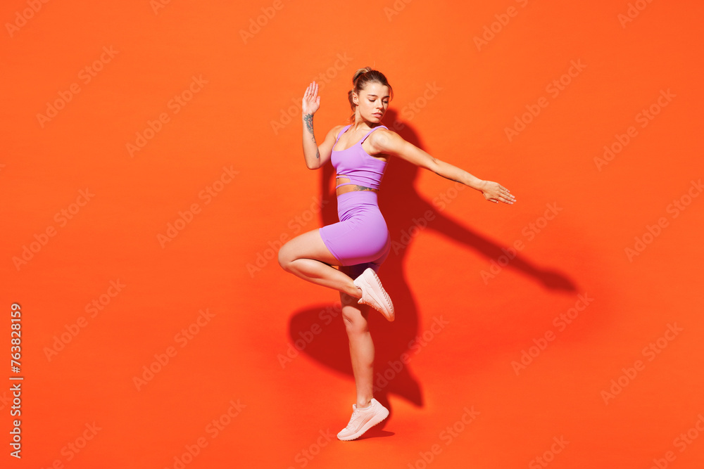 Full body side view young fitness trainer instructor woman sportsman wearing purple top clothes train in home gym raise up leg hand isolated on plain orange background. Workout sport fit abs concept.