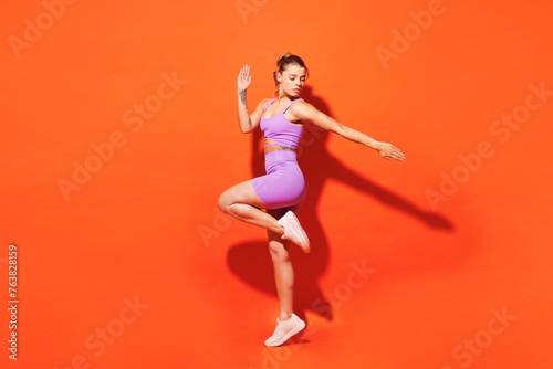 Fototapeta Naklejka Na Ścianę i Meble -  Full body side view young fitness trainer instructor woman sportsman wearing purple top clothes train in home gym raise up leg hand isolated on plain orange background. Workout sport fit abs concept.