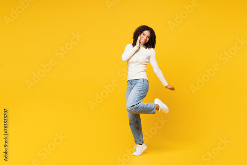 Full body little kid teen girl of African American ethnicity wear white casual clothes stand hold put hand on face raise up leg isolated on plain yellow background studio. Childhood lifestyle concept. © ViDi Studio