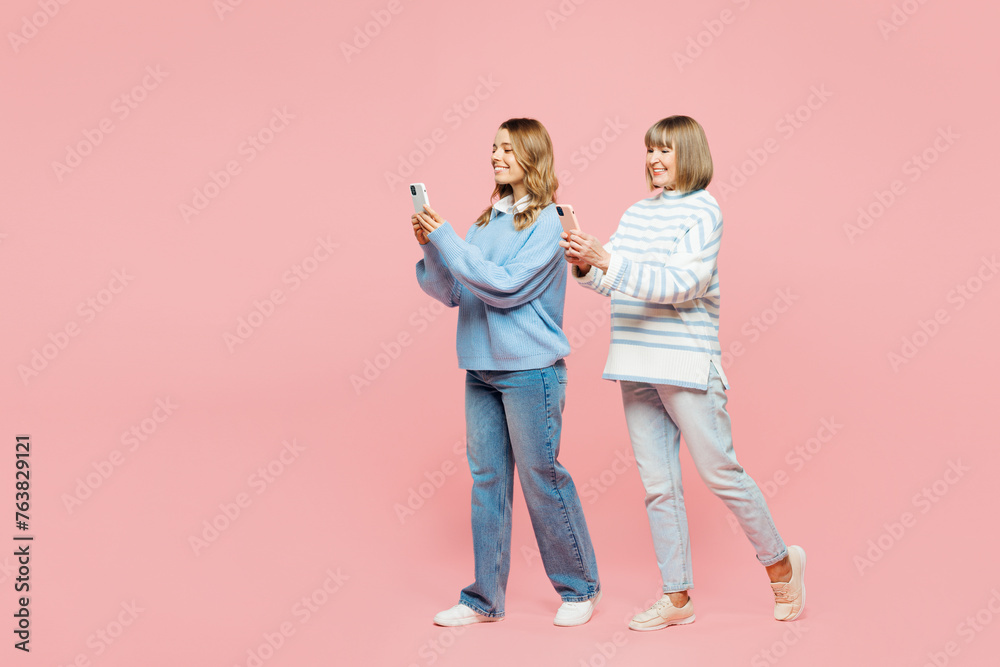 Full body elder parent mom with young adult daughter two women together wear blue casual clothes hold use mobile cell phone walk go isolated on plain pastel light pink background. Family day concept. Full body elder parent mom with young adult daughter two women together wear blue casual clothes hold use mobile cell phone walk go isolated on plain pastel light pink background. Family day concept.