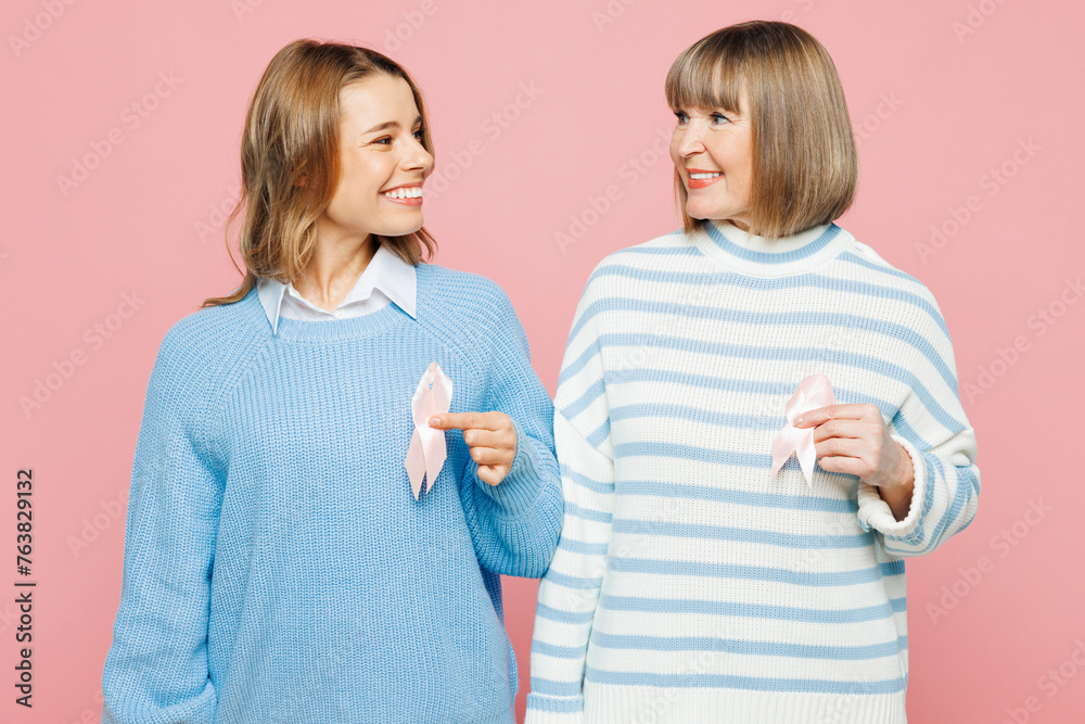 Elder fun parent mom 50s years old young adult daughter two women together wear blue casual clothes hold in hand pink ribbon symbol isolated on plain pastel light pink background. Family day concept. Elder fun parent mom 50s years old young adult daughter two women together wear blue casual clothes hold in hand pink ribbon symbol isolated on plain pastel light pink background. Family day concept.