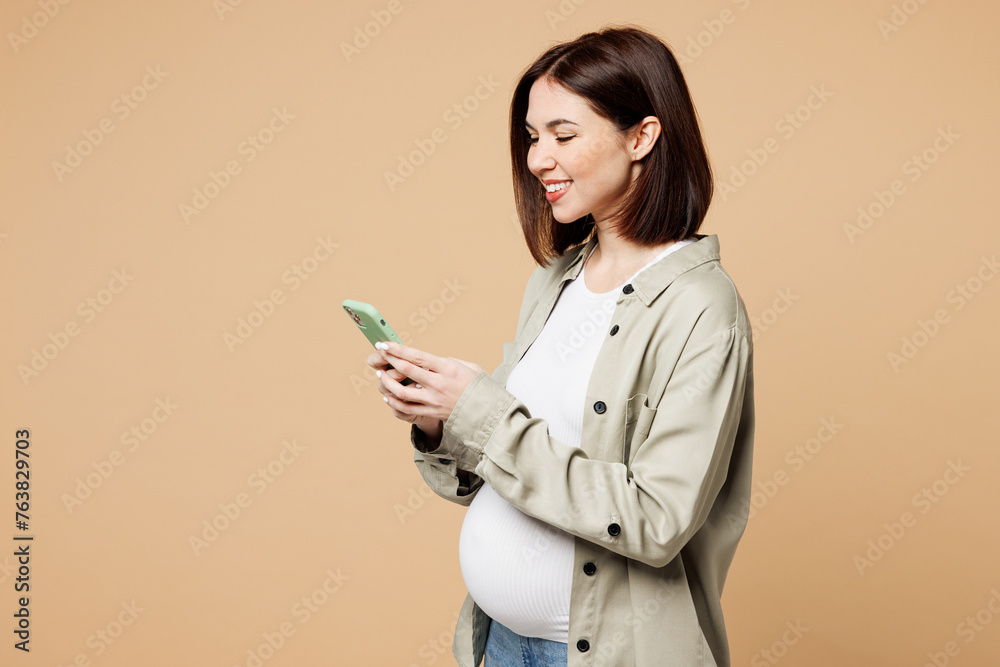 Side view young pregnant woman future mom wear grey shirt with belly stomach tummy with baby hold in hand use mobile cell phone isolated on plain beige background. Maternity family pregnancy concept. Side view young pregnant woman future mom wear grey shirt with belly stomach tummy with baby hold in hand use mobile cell phone isolated on plain beige background. Maternity family pregnancy concept.