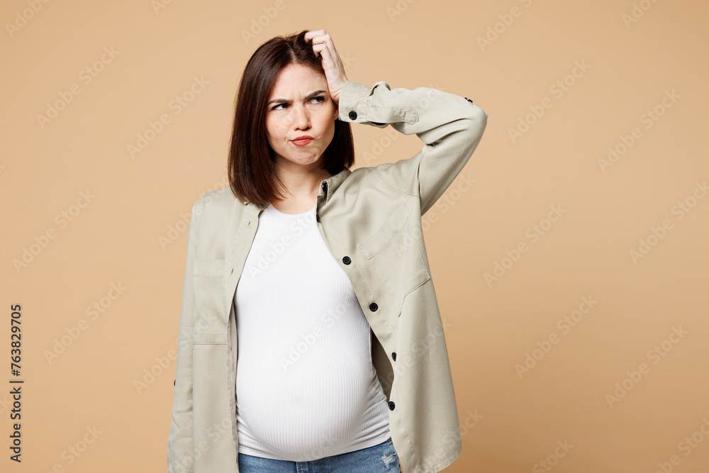 Young confused pregnant expectant woman future mom in grey shirt with belly stomach tummy with baby look aside on area hold head isolated on plain beige background Maternity family pregnancy concept