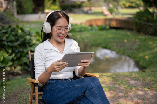 Happy Asian woman is wearing headphones and using her tablet while relaxing in her backyard garden.