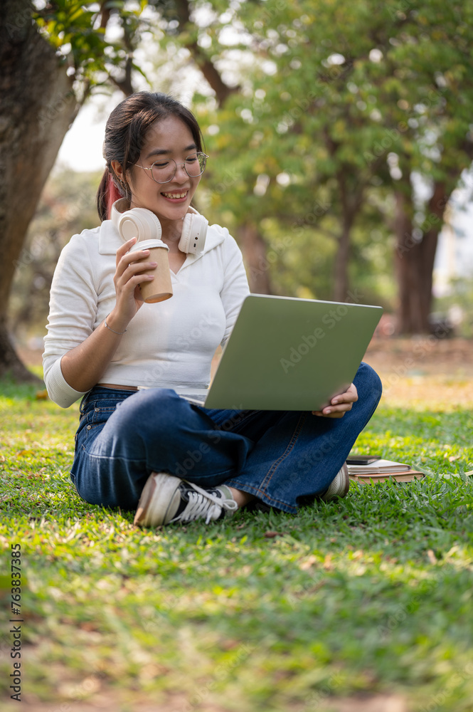 A happy young Asian woman working remotely in a green park on a bright day, using her laptop. A happy young Asian woman working remotely in a green park on a bright day, using her laptop.