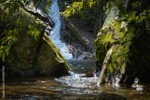 Water slide falling from the big rocks in the background, flowing between two rocks full of moss, and sunlight shines on the forest which covered the river, in Nuandong valley, Keelung city, Taiwan.