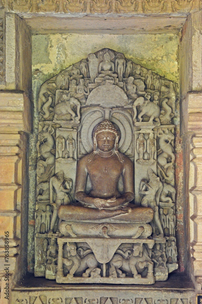 A finely detailed stone sculpture of a  Jain Tirthankara ADINATH,  with a serene expression, surrounded by ornate carvings 