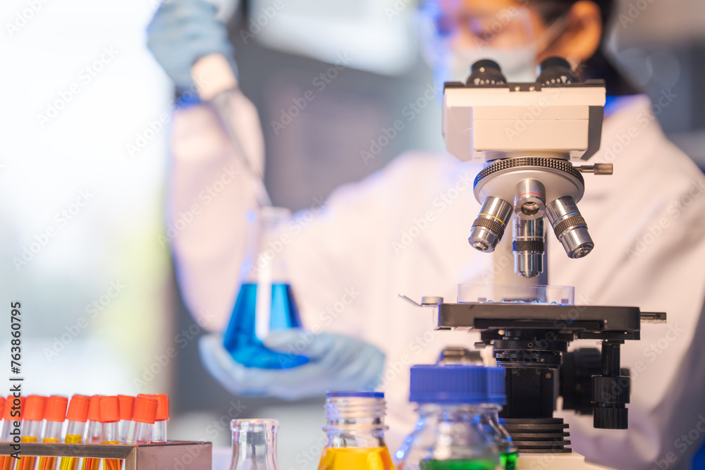 Close up female scientist in white lab coat working in biotechnological laboratory, using microscope, pipette, glass flask in clinical laboratory. Close up female scientist in white lab coat working in biotechnological laboratory, using microscope, pipette, glass flask in clinical laboratory.