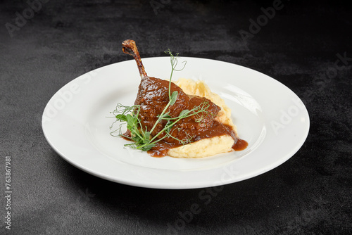 Classic dish french cuisine - roasted duck leg in sauce with garnish on black background. Cooked duck leg with mash potatoes on white plate on dark concrete table. Duck confit in minimal style. photo