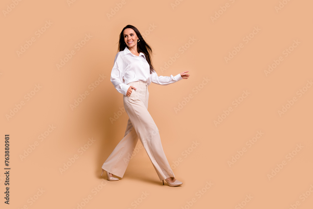 Full length photo of positive dreamy woman wear white shirt carefree walking empty space isolated beige color background