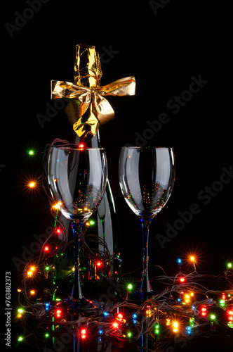 A closed bottle of champagne and two glasses with a multicolored electric garland on a black background