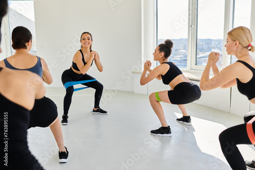 group of happy diverse women in sportswear looking at trainer and squatting with resistance bands