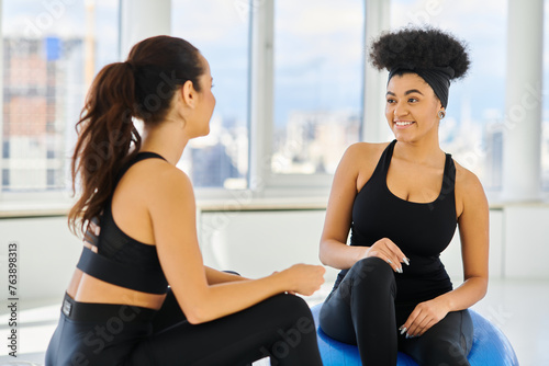 happy and diverse female friends sitting on fitness balls and smiling during pilates class