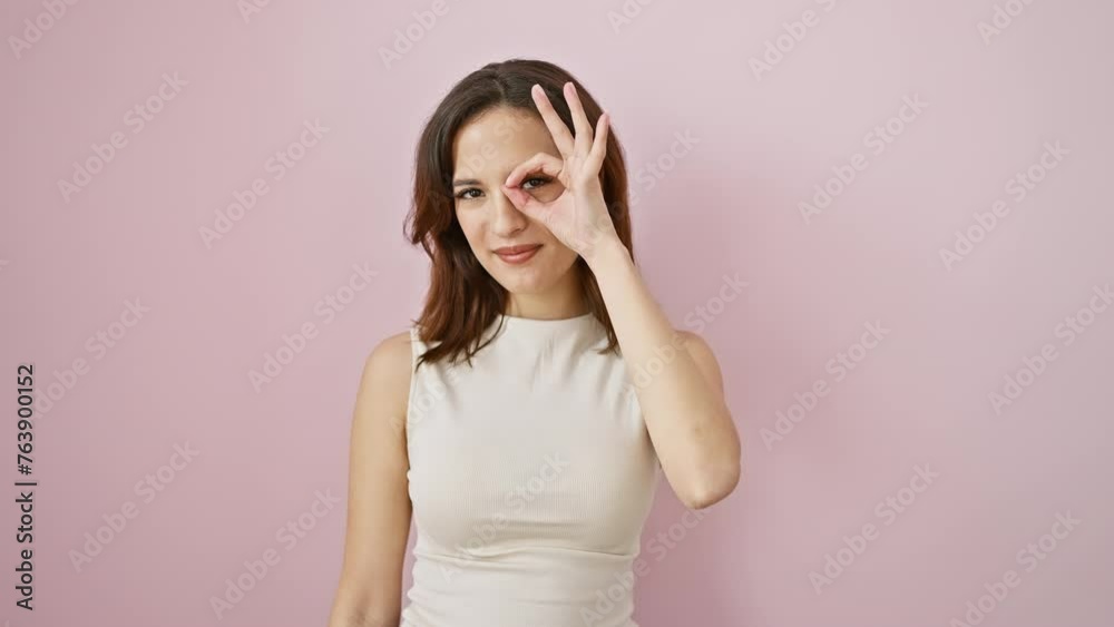 Cheerful young hispanic woman strikes a fun 'ok' hand gesture, peeking through her fingers. standing, sporting a comfy sleeveless tee, against a vibrant isolated pink backdrop. Cheerful young hispanic woman strikes a fun 'ok' hand gesture, peeking through her fingers. standing, sporting a comfy sleeveless tee, against a vibrant isolated pink backdrop.