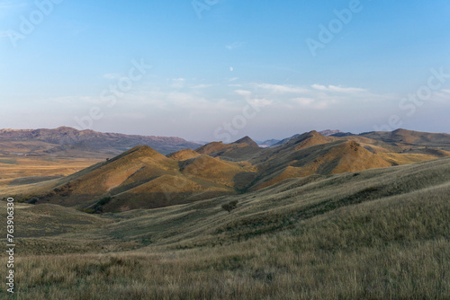 Mountain savannah landscape. Mountain peaks are visible. Dry green and orange grass. Light blue sky with clouds.