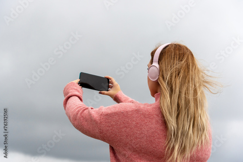 Long-haired blonde woman with her back turned, wearing a pink jumper and large matching headphones, listening to music by the sea and taking a picture of the landscape on a cloudy day.