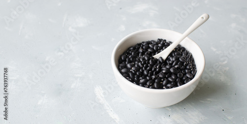 Raw black bens in a white bowl with a teaspoon on a gray background. Concept of healthy eating. Vegetarian and vegan food. Horizontal orientation. Selective focus. Bunner