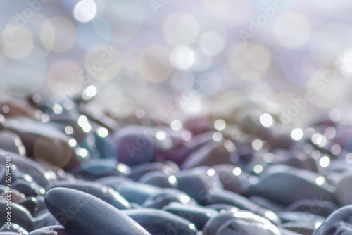 Pebble stones on sea shore close up in defocused multicolored bokeh lights. Grey pebble stones background, soft focus.