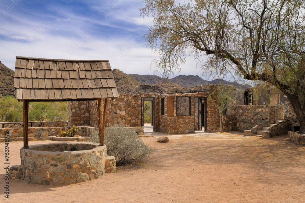 A historic stone building with a wooden roof stands deserted among desert flora under a bright blue sky