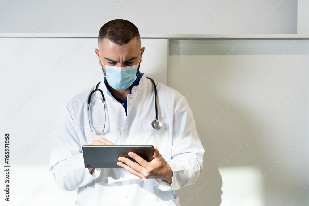 Male doctor wearing medical mask and holding tablet computer in hospital setting, appearing vigilant and prepared to tend to patients with utmost expertise and care. Male doctor wearing medical mask and holding tablet computer in hospital setting, appearing vigilant and prepared to tend to patients with utmost expertise and care.