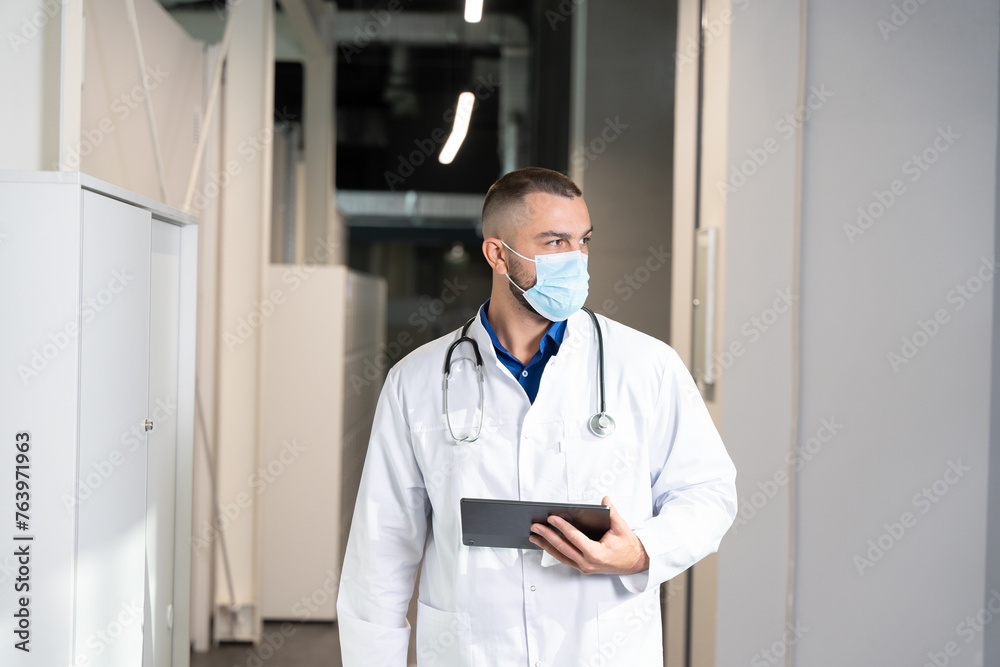Professional male doctor wearing a medical mask and holding a digital tablet computer walking through the hospital corridor. Healthcare professional on duty in a busy hospital setting. Professional male doctor wearing a medical mask and holding a digital tablet computer walking through the hospital corridor. Healthcare professional on duty in a busy hospital setting.