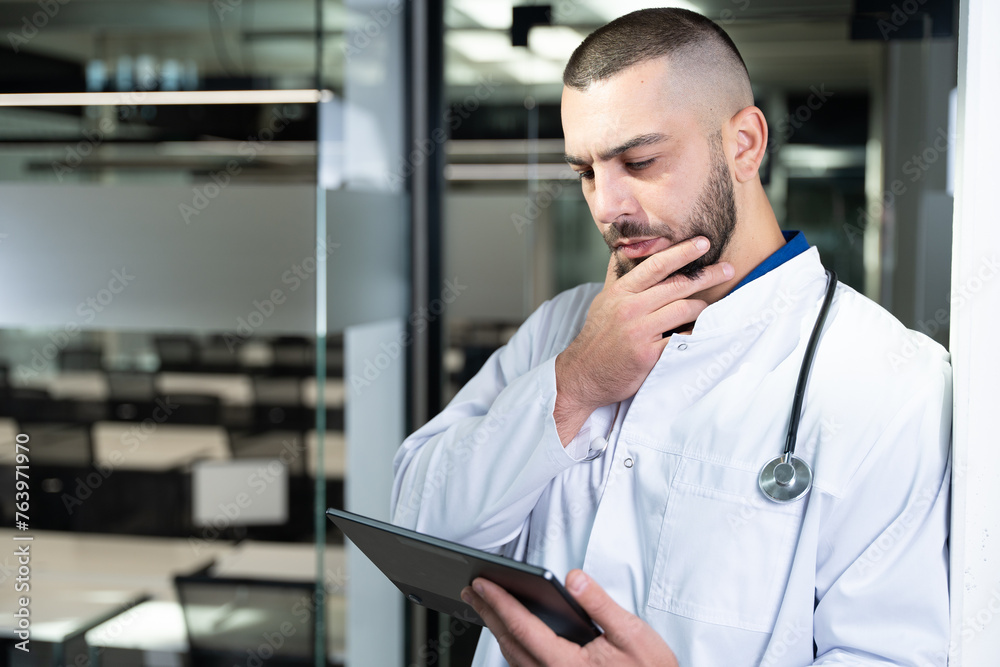 Concerned male doctor in medical attire is holding and looking at tablet computer with worried expression, standing in a modern medical facility. Technology and healthcare concepts. Concerned male doctor in medical attire is holding and looking at tablet computer with worried expression, standing in a modern medical facility. Technology and healthcare concepts.
