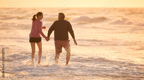 Loving Retired Senior Couple On Vacation Walking Along Beach Through Waves Holding Hands At Sunrise