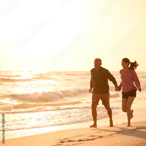 Loving Retired Senior Couple On Vacation Running Along Beach Shoreline Holding Hands At Sunrise