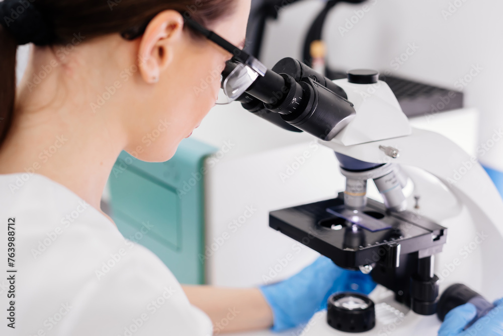 In a modern medical laboratory: a microbiologist in glasses looks under a microscope, analyzing a sample. Scientist working with high-tech equipment. Close-up In a modern medical laboratory: a microbiologist in glasses looks under a microscope, analyzing a sample. Scientist working with high-tech equipment. Close-up