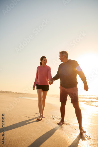 Loving Retired Senior Couple On Vacation Walking Along Beach Shoreline Holding Hands At Sunrise