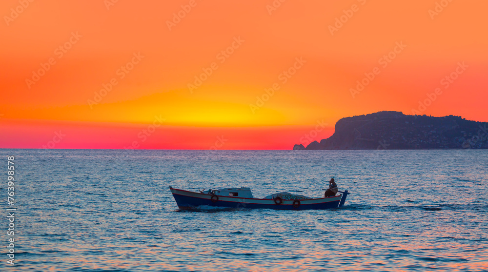 A fisherman departs Alanya Harbor on the Mediterranean sea in Alanya peninsula in the background at dusk - Alanya, Turkey 