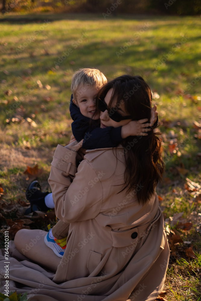Fototapeta premium A mother and son embrace and play in the spring park. The boy holds green tree leaves while the mother tickles him, creating precious family moments on Mother's Day, enjoying a picnic in nature.
