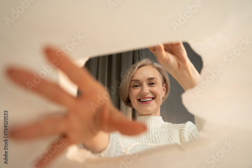 A joyful woman is captured looking inside a cardboard box, as she appears to be in the process of unpacking or discovering items during a move or relocation. The shape of the box opening frames her photo