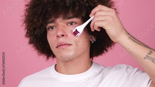 A young man with full, visible tattoos on his arm holds a derma roller to his cheek against a pink background.