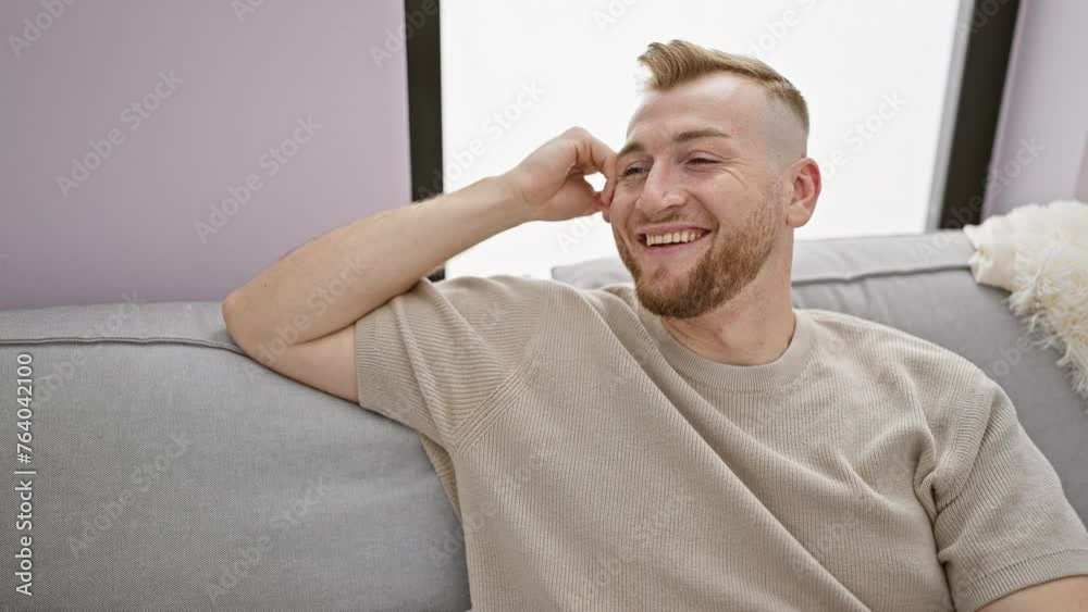 A cheerful bearded young man relaxing on a sofa indoors with a neutral modern decor expressing happiness and comfort. A cheerful bearded young man relaxing on a sofa indoors with a neutral modern decor expressing happiness and comfort.