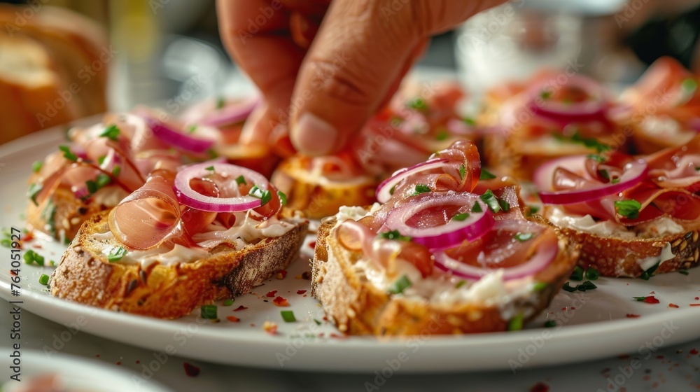 Hand garnishing open-faced ciabatta sandwiches with prosciutto, cream cheese, red onion, and chives on a white plate