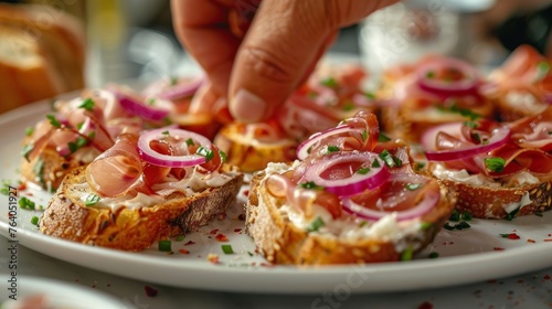 Hand garnishing open-faced ciabatta sandwiches with prosciutto, cream cheese, red onion, and chives on a white plate